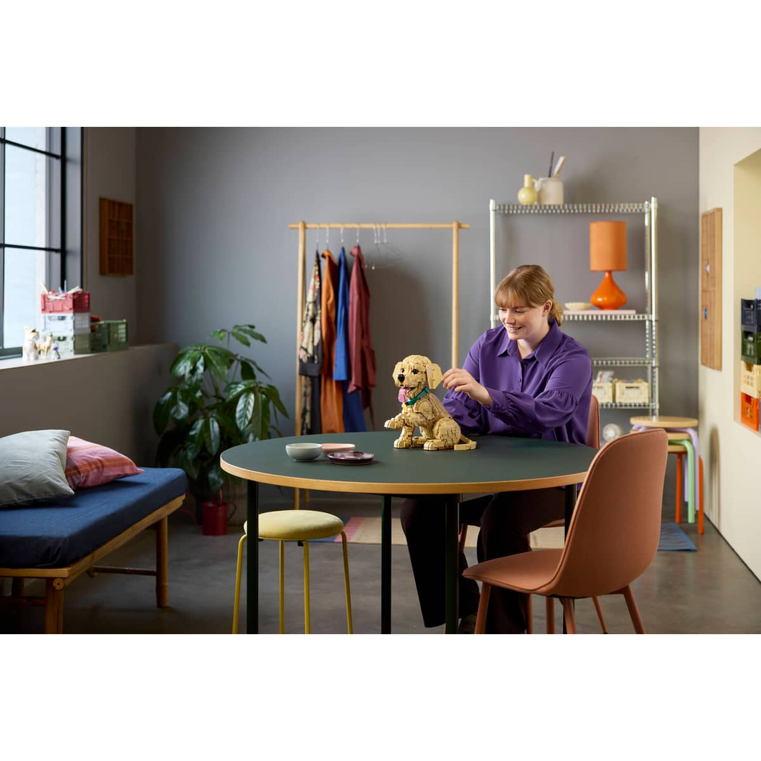 Een vrouw zit aan tafel en bouwt een Lego-hond. Een kledingrek staat in de buurt.