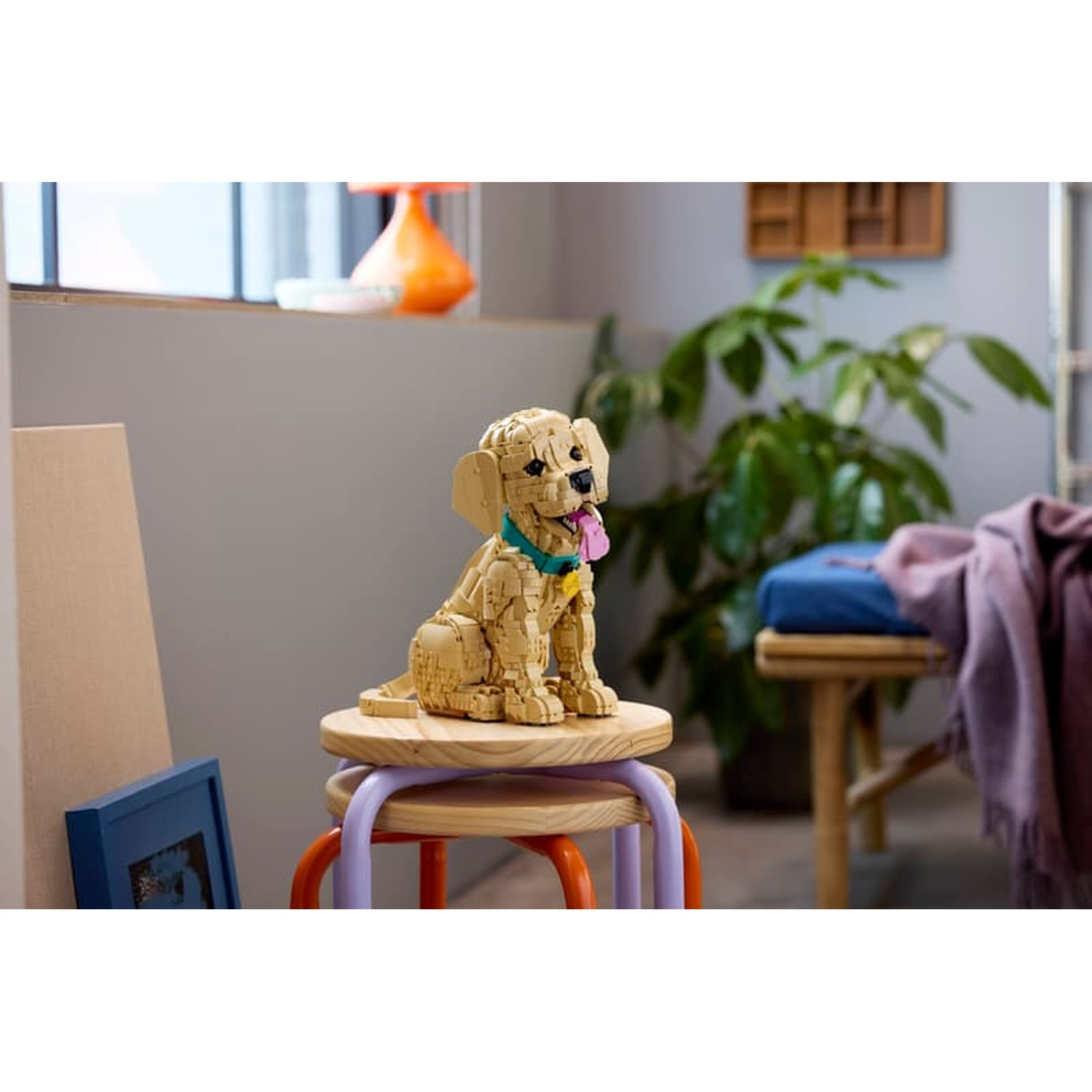 Een Lego-hond zit op een krukje, met een fotolijst en plant in de buurt.