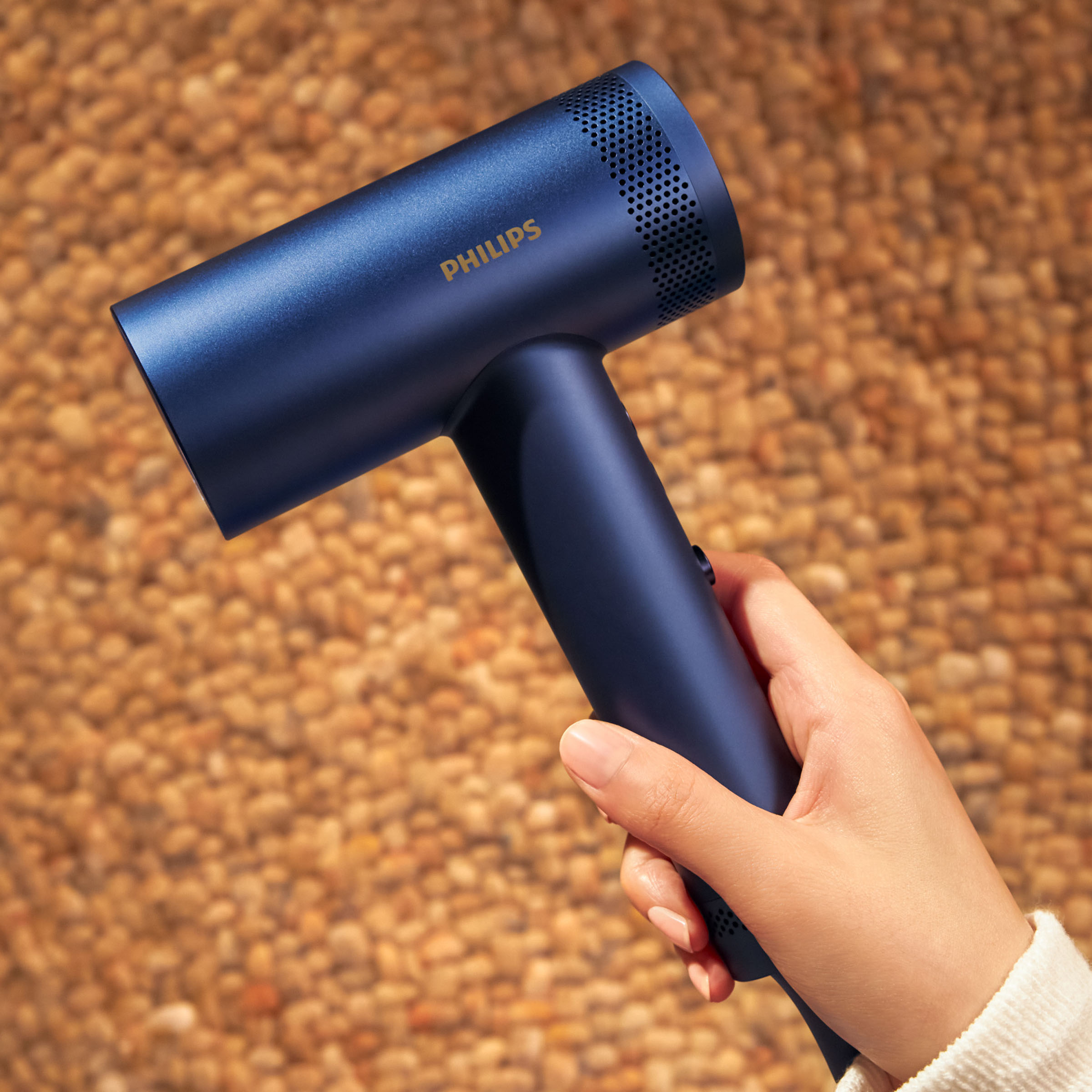 Niebieska suszarka do włosów Philips jest trzymana przez dłoń na tle kamyków.