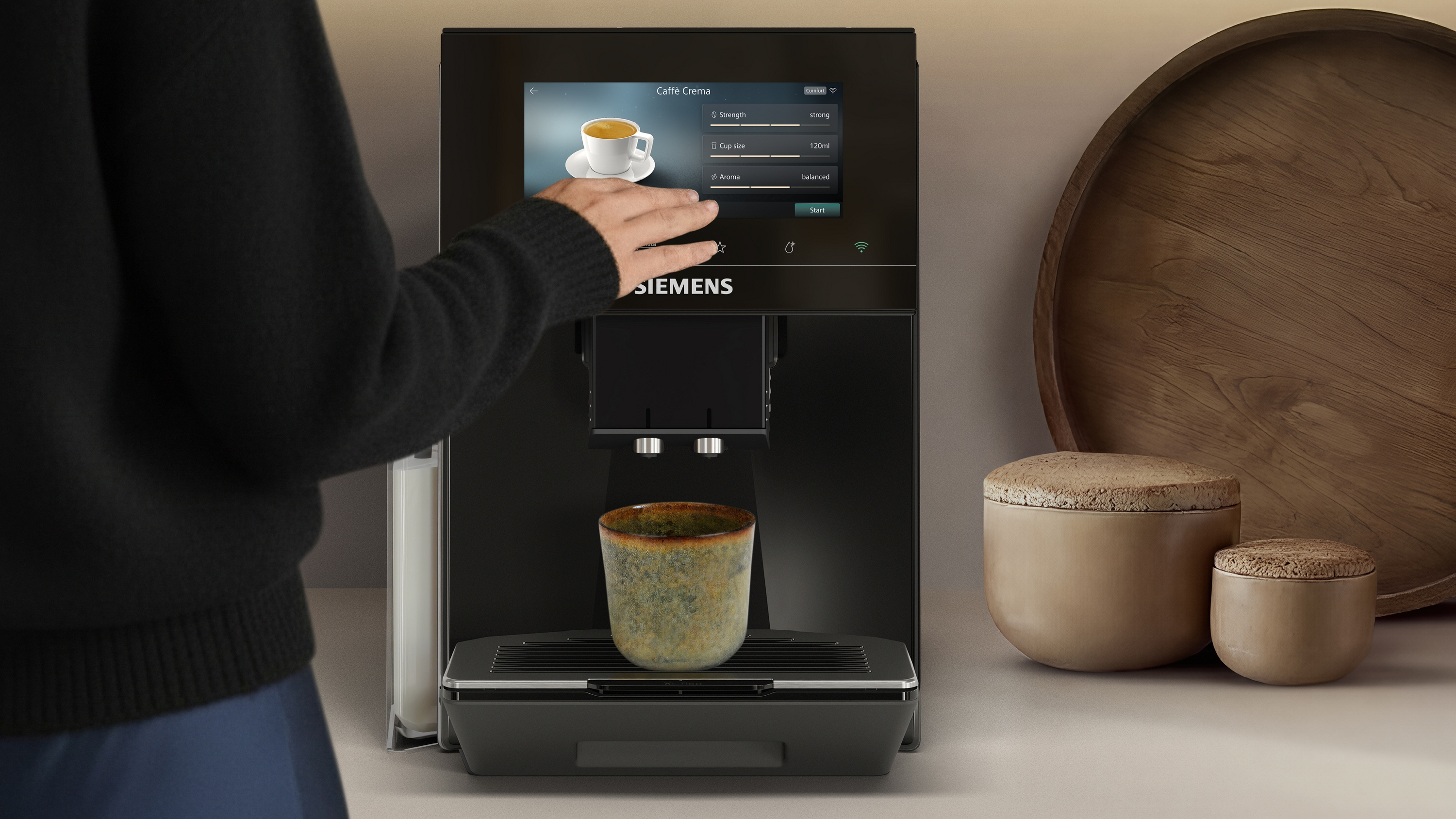 Une main utilise une machine à café avec écran tactile. Une tasse est prête.