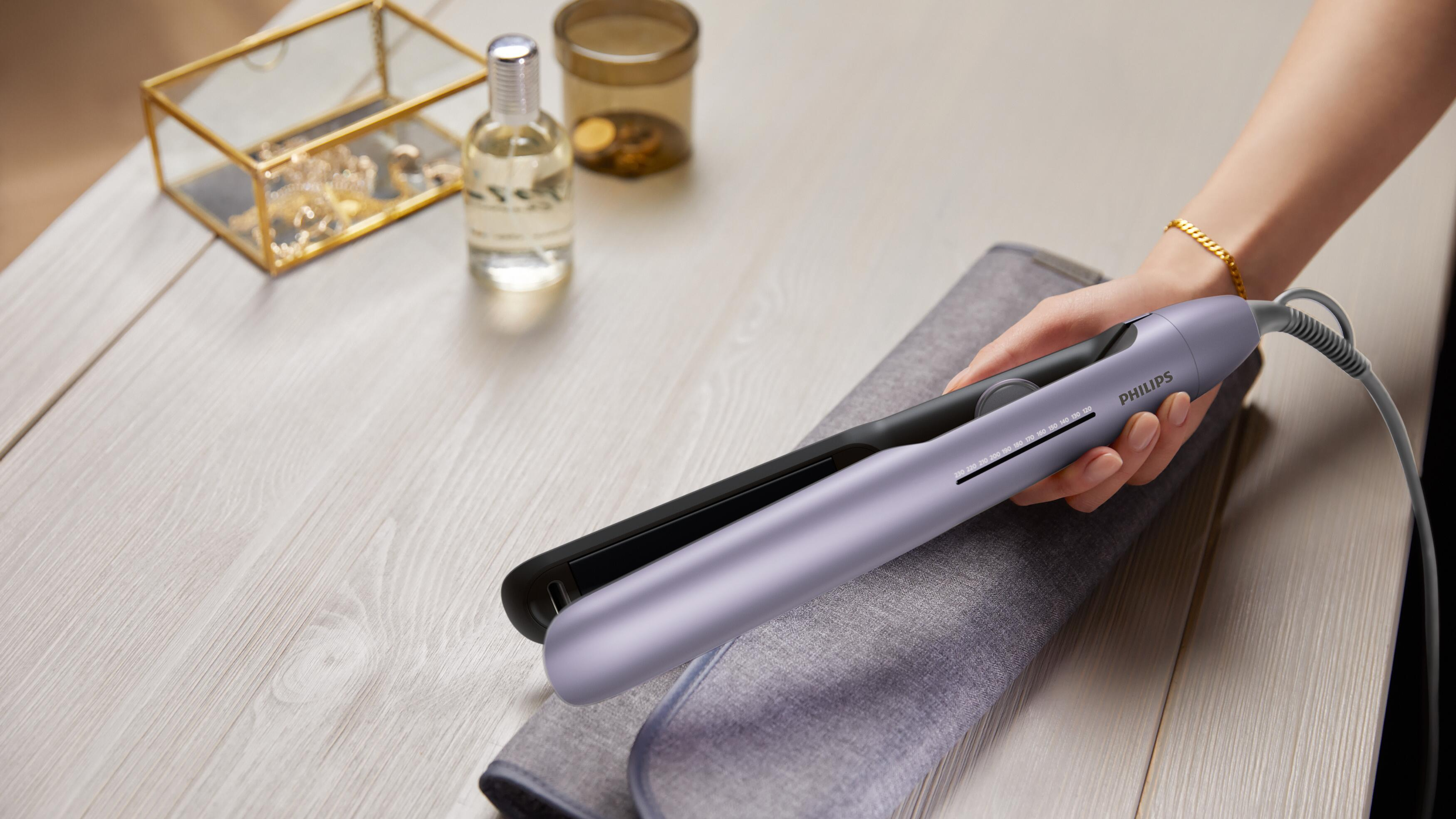 Ręka trzyma fioletową prostownicę Philips na szarym materiale na drewnianym stole.