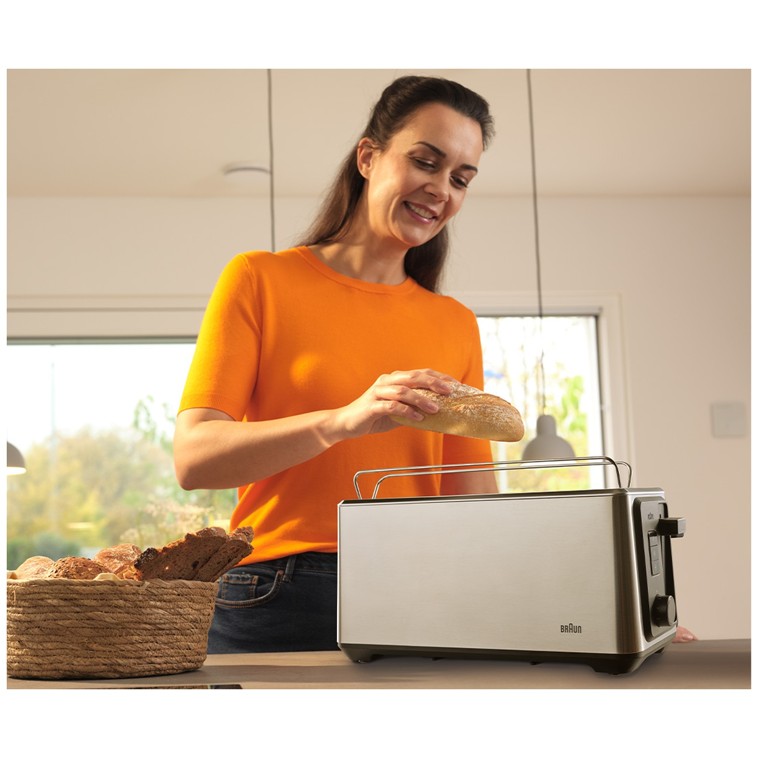 A woman in an orange shirt puts a bread roll into a toaster. Next to it, a basket of bread.