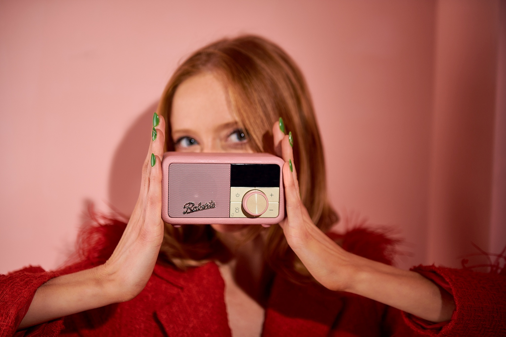 Une femme aux cheveux roux et aux ongles verts tient une radio rose devant son visage.