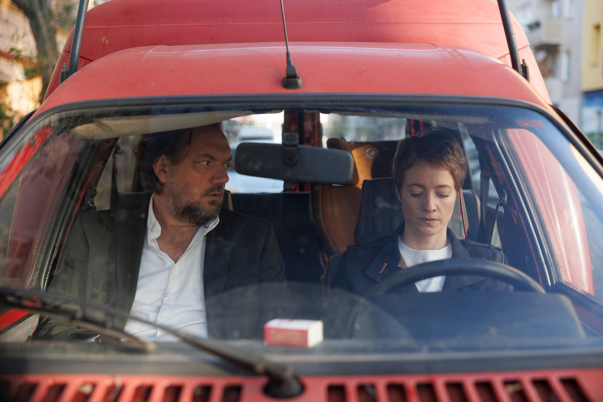 Mann und Frau sitzen in einem roten Auto. Sie schauen nachdenklich.