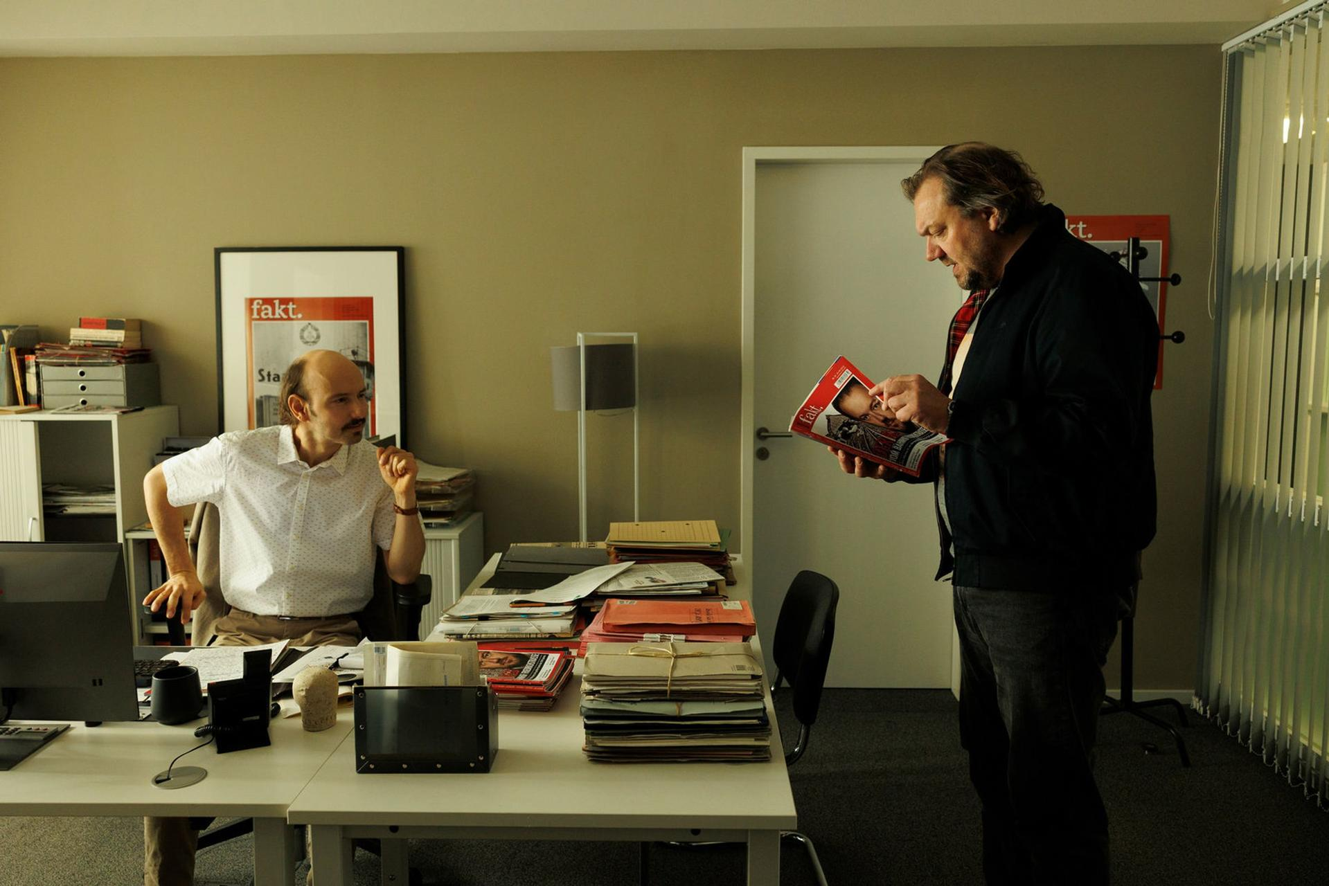 Ein Mann sitzt am Schreibtisch, ein anderer steht und liest ein Magazin.