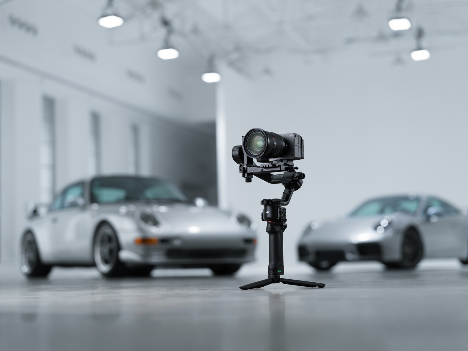 Un gimbal con cámara en primer plano, dos coches plateados al fondo.