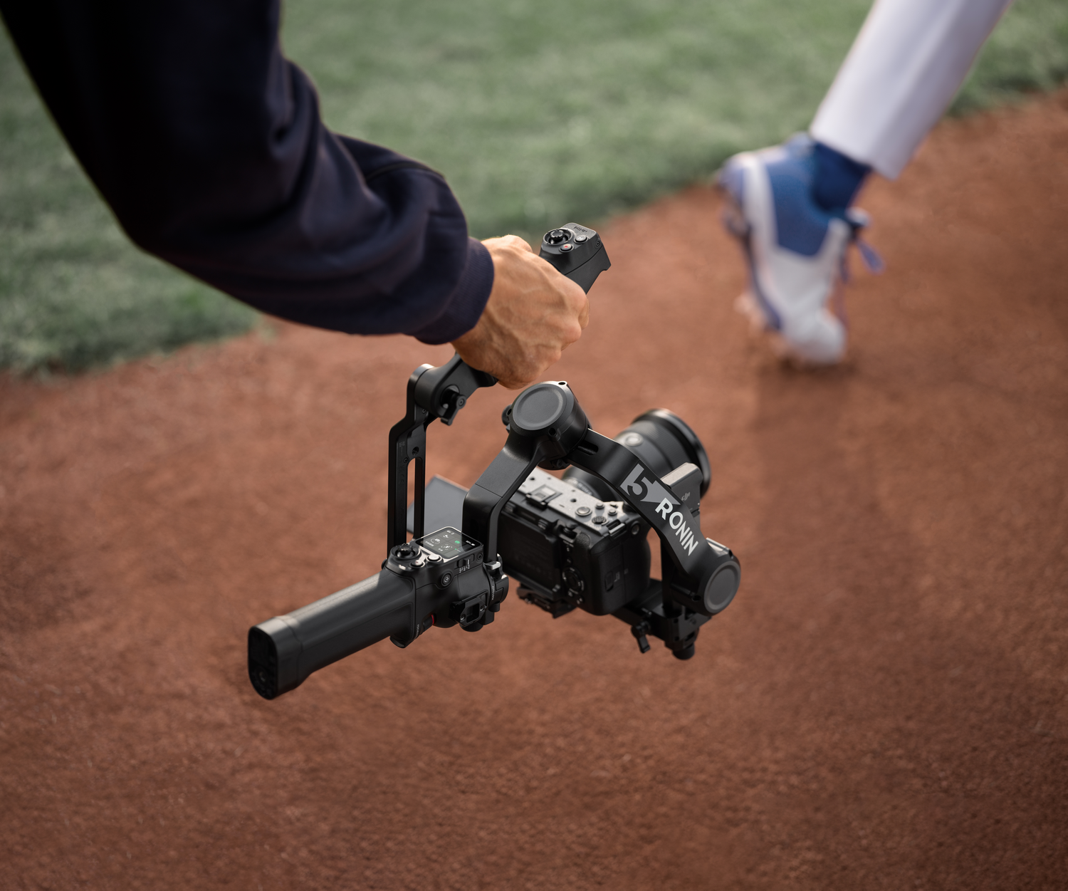 Una mano sostiene un gimbal con cámara en un campo de béisbol, un jugador al fondo.