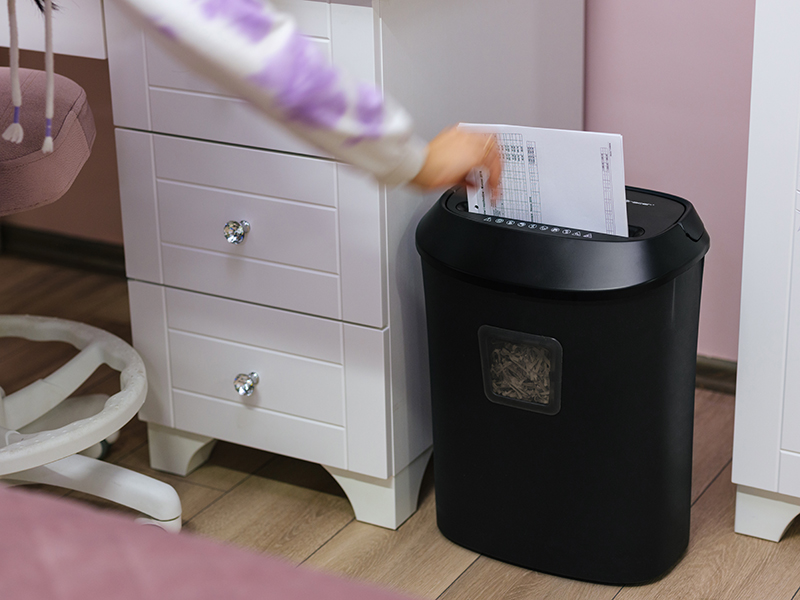 Ręka niszczy papier w czarnej niszczarce obok białej komody.