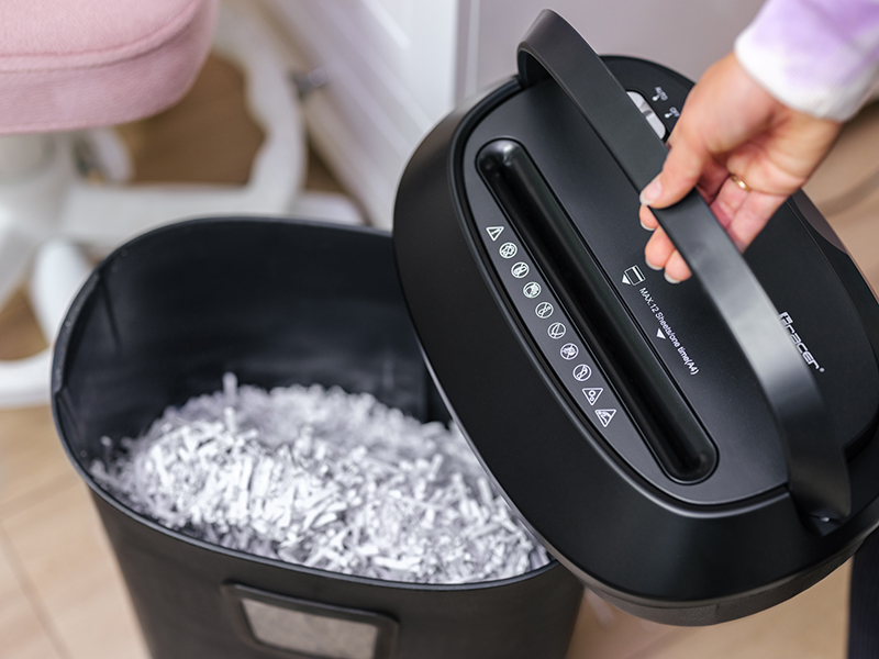 Dłoń trzyma uchwyt czarnej niszczarki do papieru nad koszem pełnym pociętego papieru.