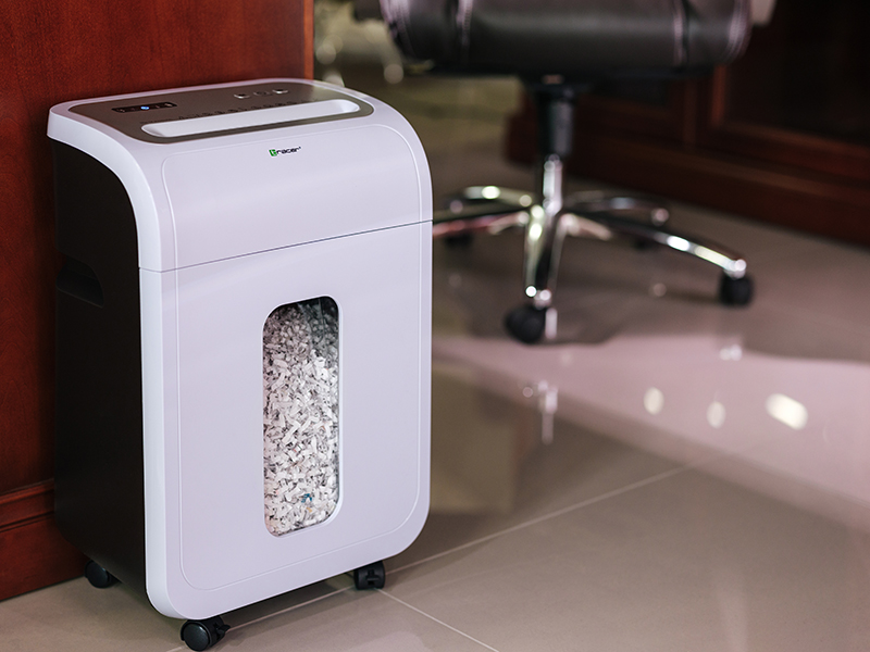 Biała niszczarka do papieru z kółkami stoi obok drewnianej szafki. Krzesło biurowe w tle.