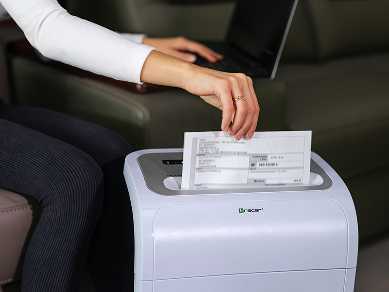 Ręka wkłada dokument do niszczarki, podczas gdy osoba pracuje na laptopie.