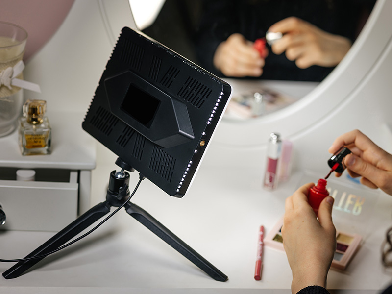 Osoba nakłada lakier do paznokci, obok lampa pierścieniowa na statywie.