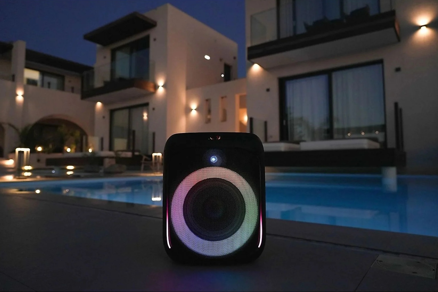 Czarny głośnik z kolorowymi światłami stoi nocą przy basenie przed domem.