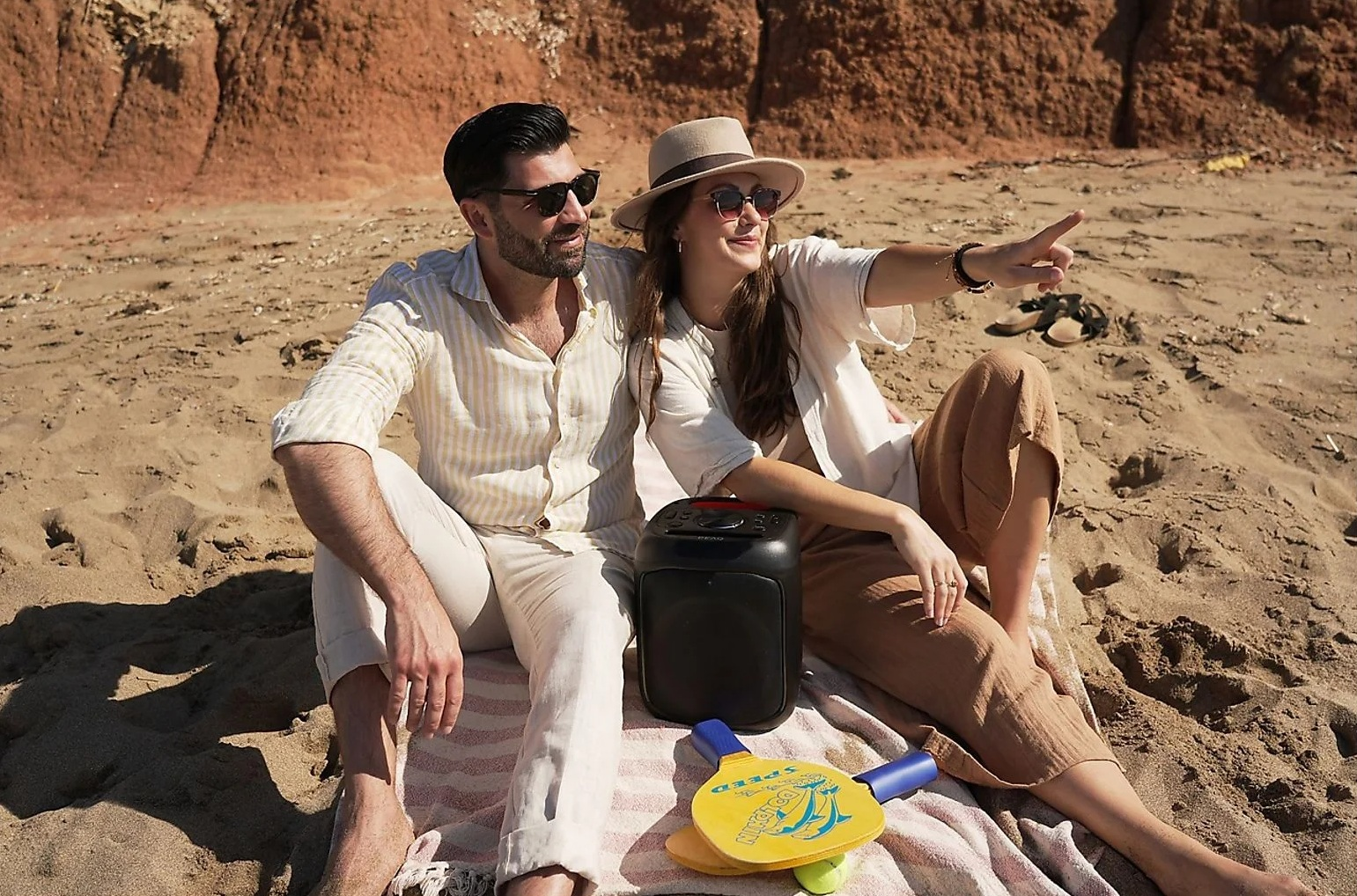 Mężczyzna i kobieta siedzą na piaszczystej plaży z głośnikiem i zabawkami do paddle.