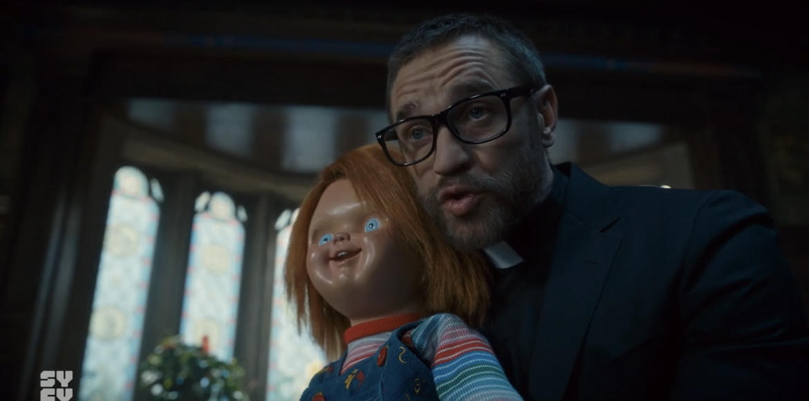 Ein Mann mit Brille und Priesterkragen hält Chucky, die Puppe, im Arm.