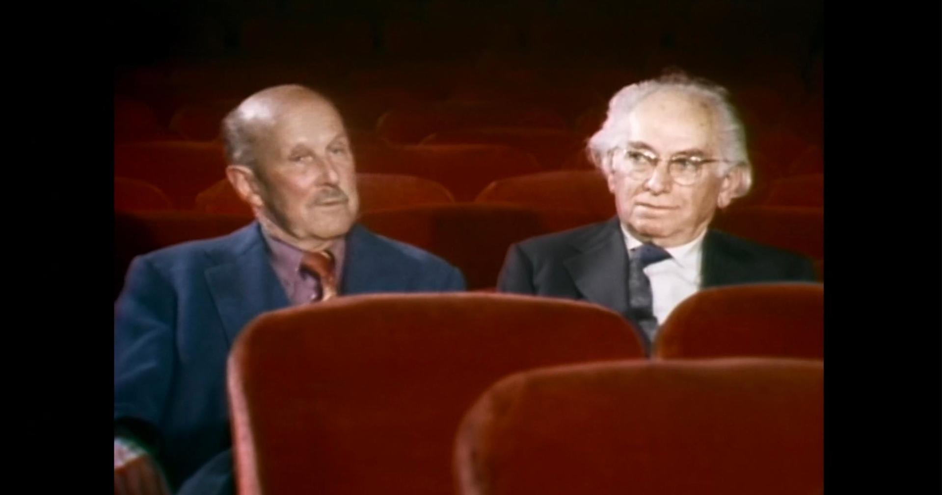 Zwei ältere Männer in einem Kino mit roten Sitzen.