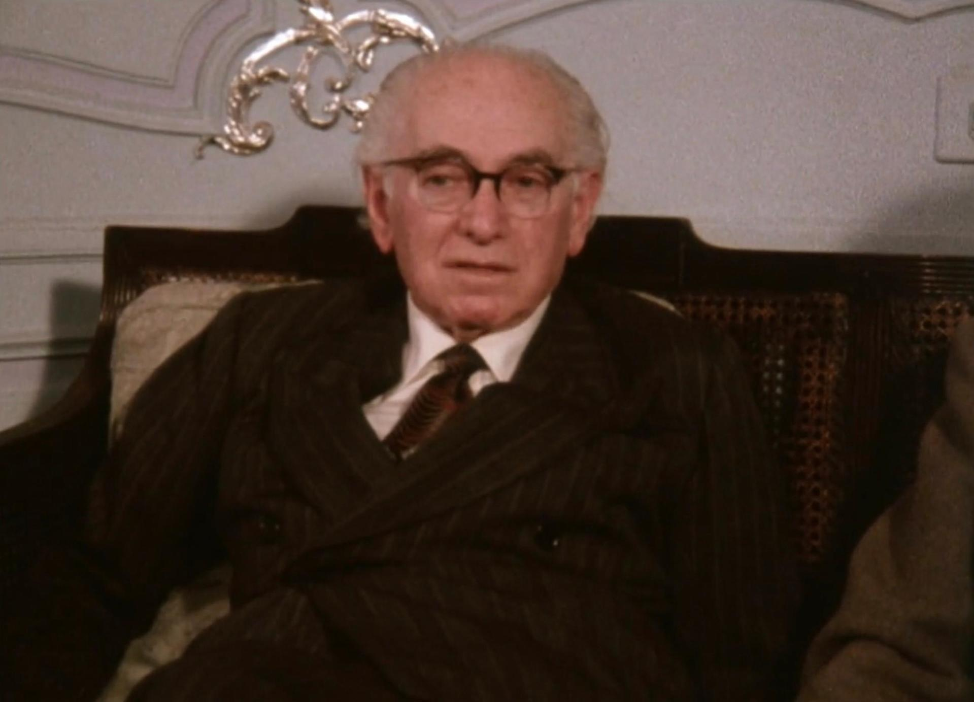 Ein älterer Mann mit Brille sitzt auf einer Couch. Er trägt einen Anzug.