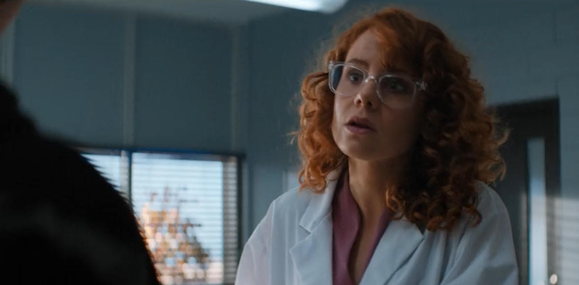 Eine Frau mit roten Locken und Brille trägt einen weißen Laborkittel.