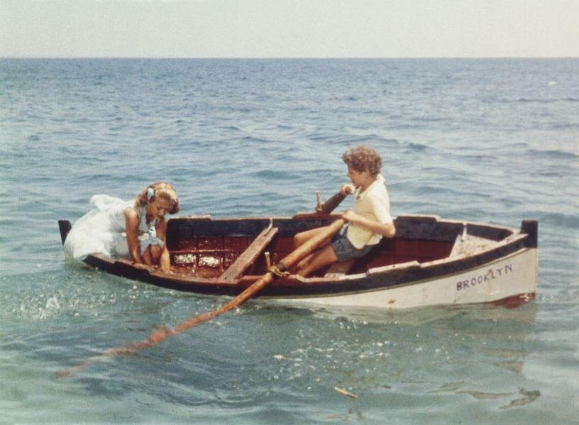 Zwei Kinder in einem Ruderboot auf dem Meer. Ein Junge rudert, ein Mädchen sitzt vorne.