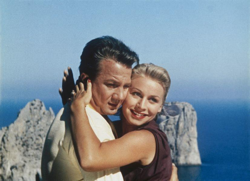 Ein Mann und eine Frau umarmen sich vor felsiger Küste und blauem Himmel.