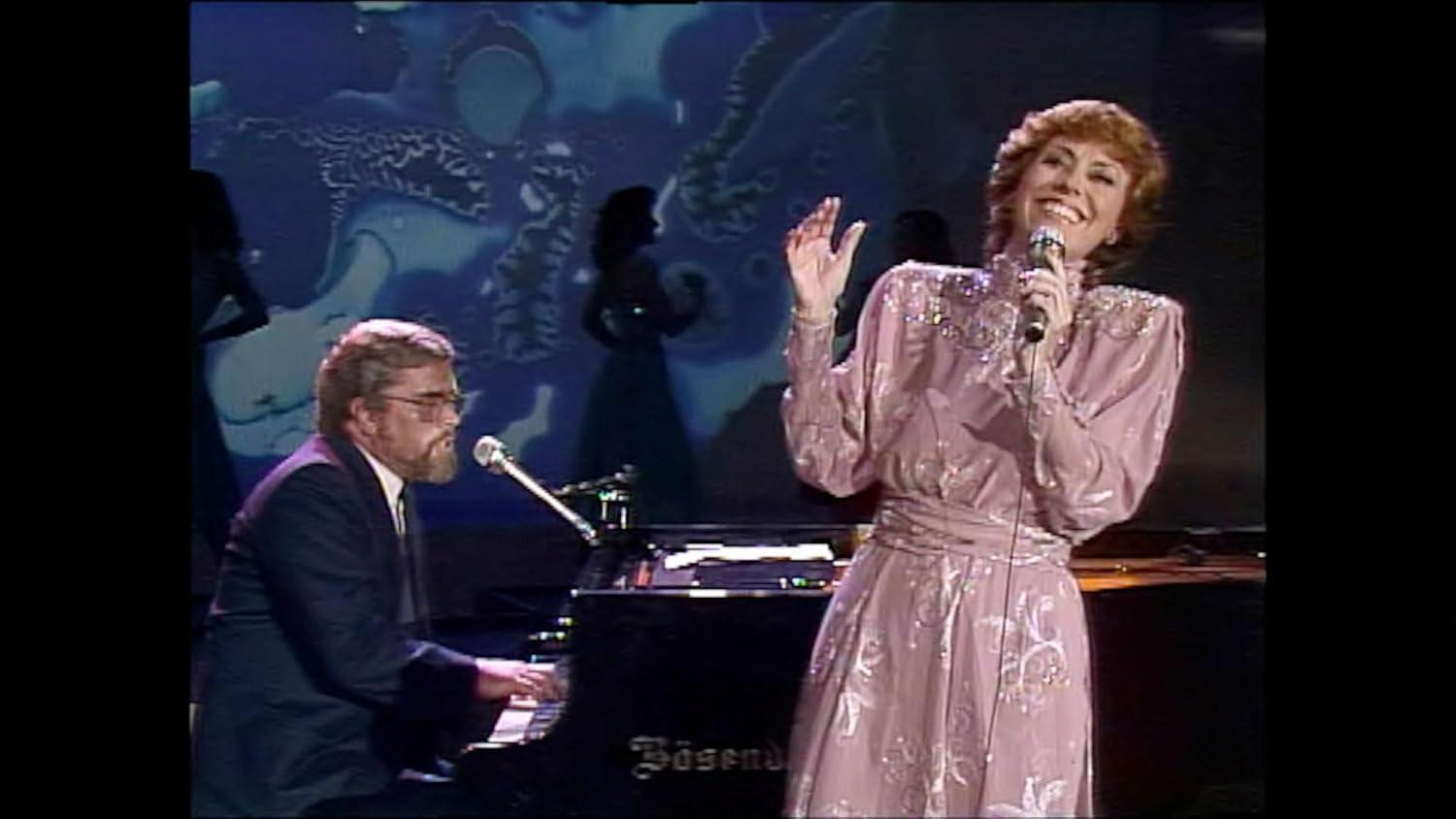 Eine Sängerin in einem rosa Kleid singt ins Mikrofon. Ein Mann spielt Klavier.
