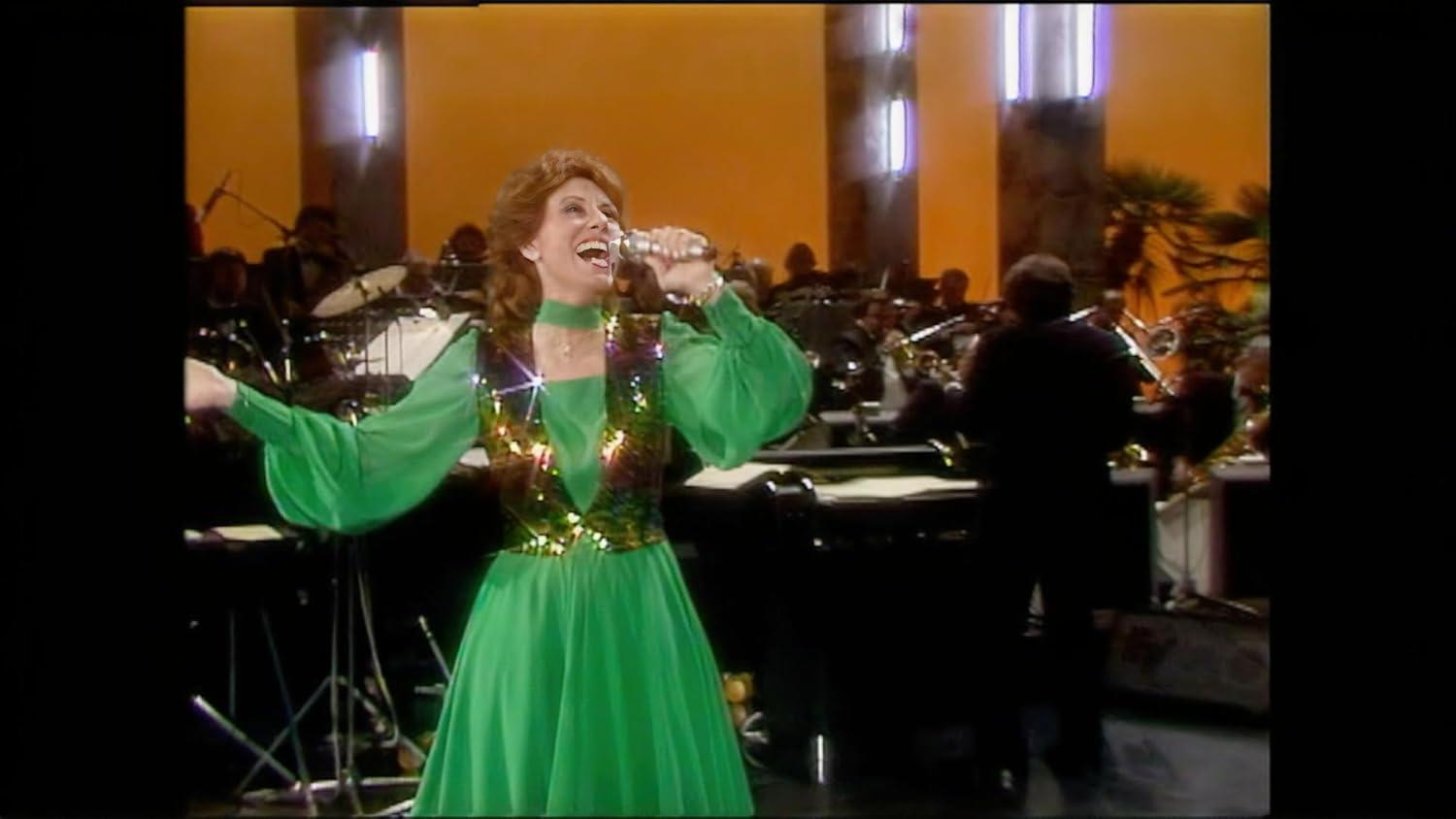 Sängerin in grünem Kleid mit Glitzerweste singt ins Mikrofon, Orchester im Hintergrund.
