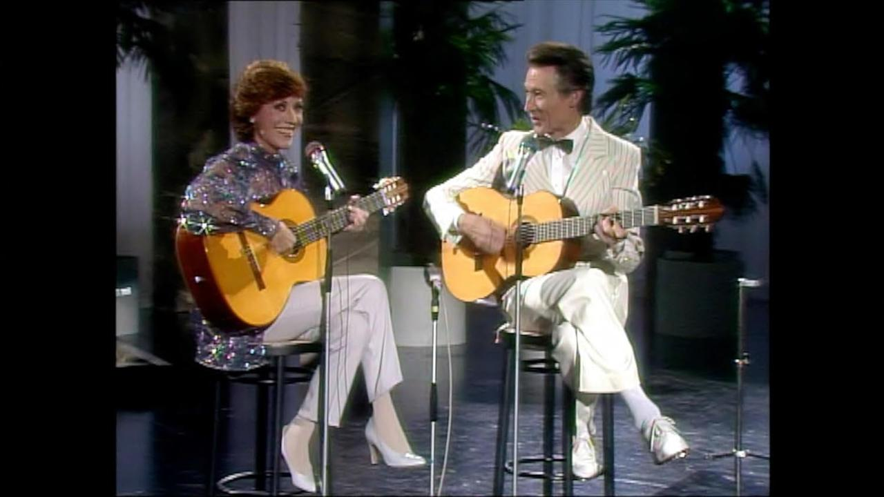 Eine Frau und ein Mann sitzen auf Hockern und spielen Gitarre.