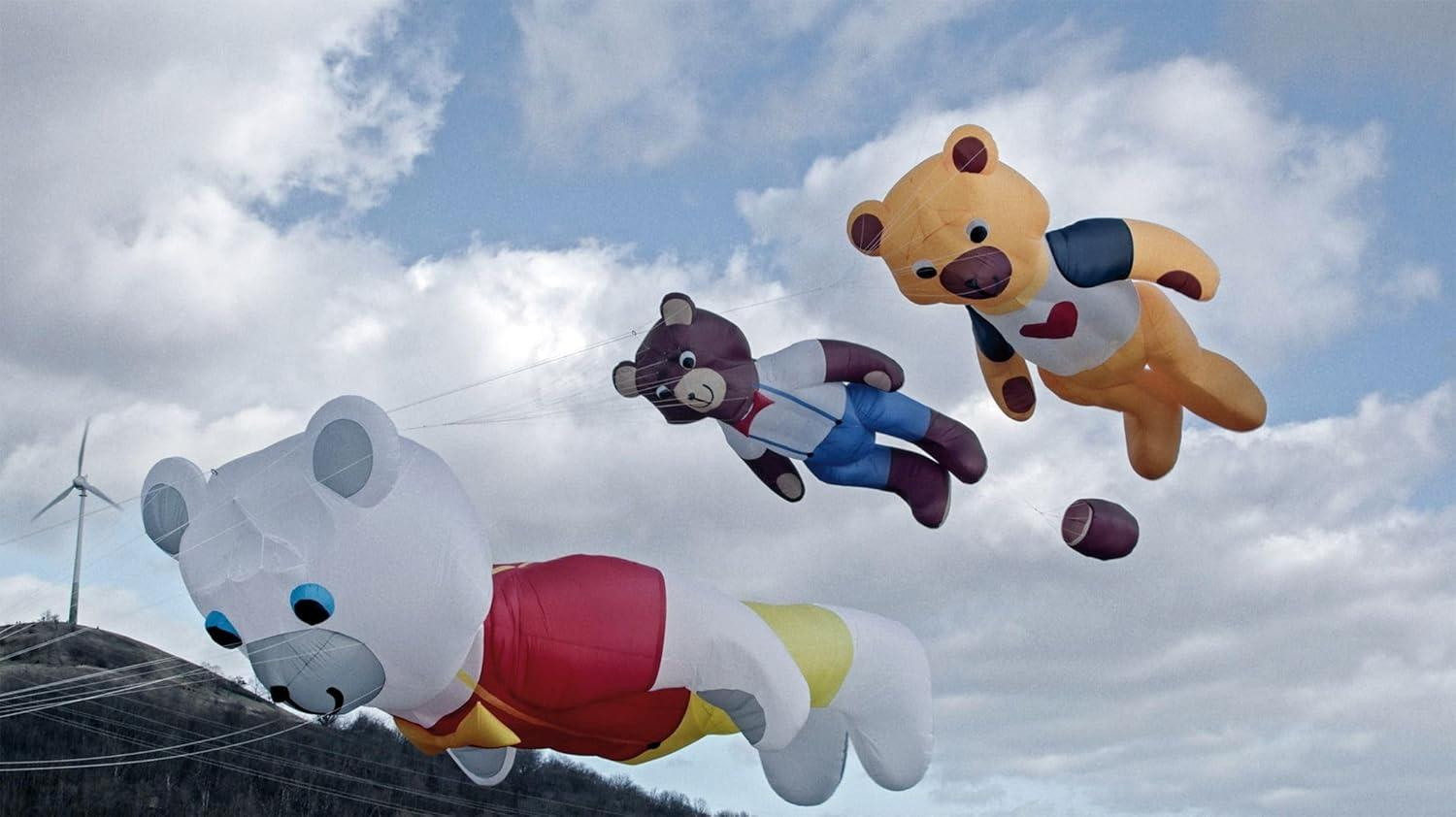 Drei aufblasbare Teddybären schweben am bewölkten Himmel neben einer Windturbine auf einem Hügel.