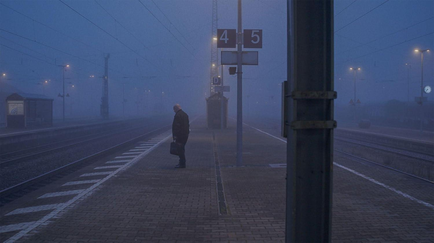 Ein Mann mit Koffer wartet im Nebel auf einem Bahnsteig neben den Gleisen.
