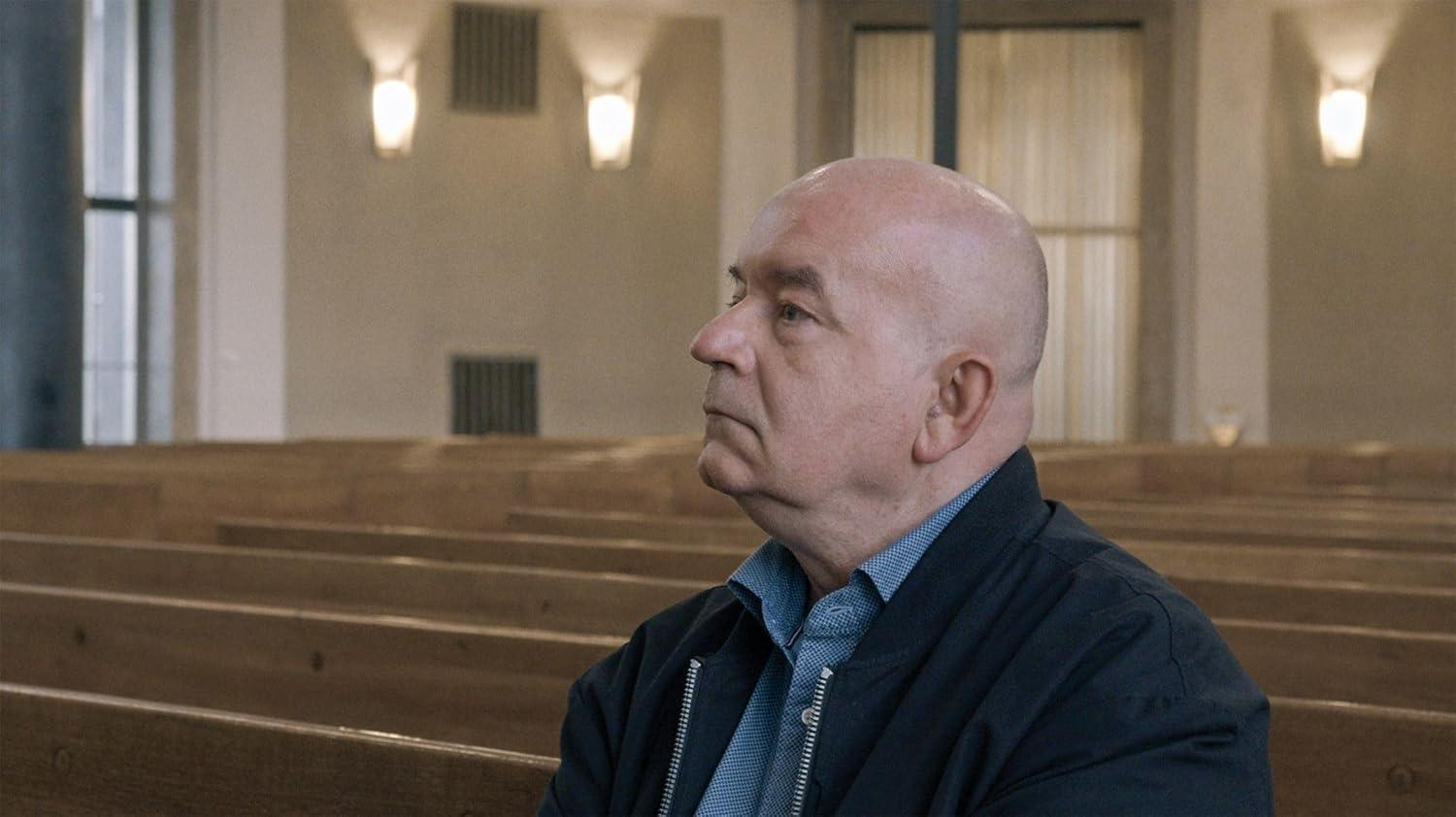 Ein Mann mit Glatze sitzt in einer Holzkirche und blickt nach rechts.