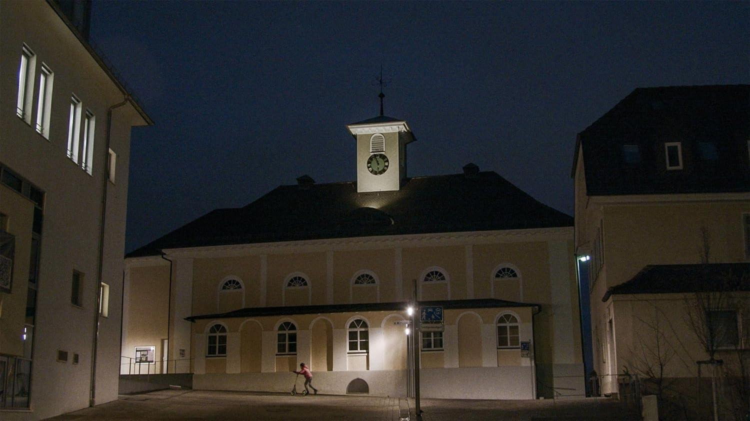 Ein Kind fährt Roller vor einem beleuchteten Gebäude mit einem Uhrturm bei Nacht.