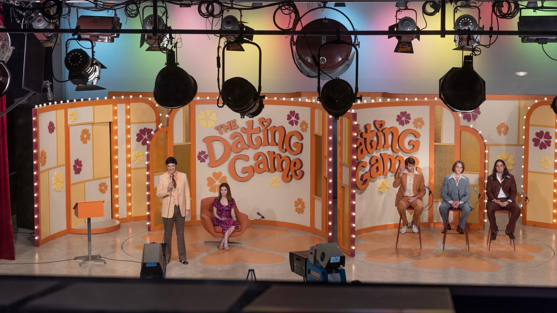 Eine Fernsehsendung mit dem Titel „The Dating Game“. Ein Moderator steht neben einer Frau, drei Männer sitzen.