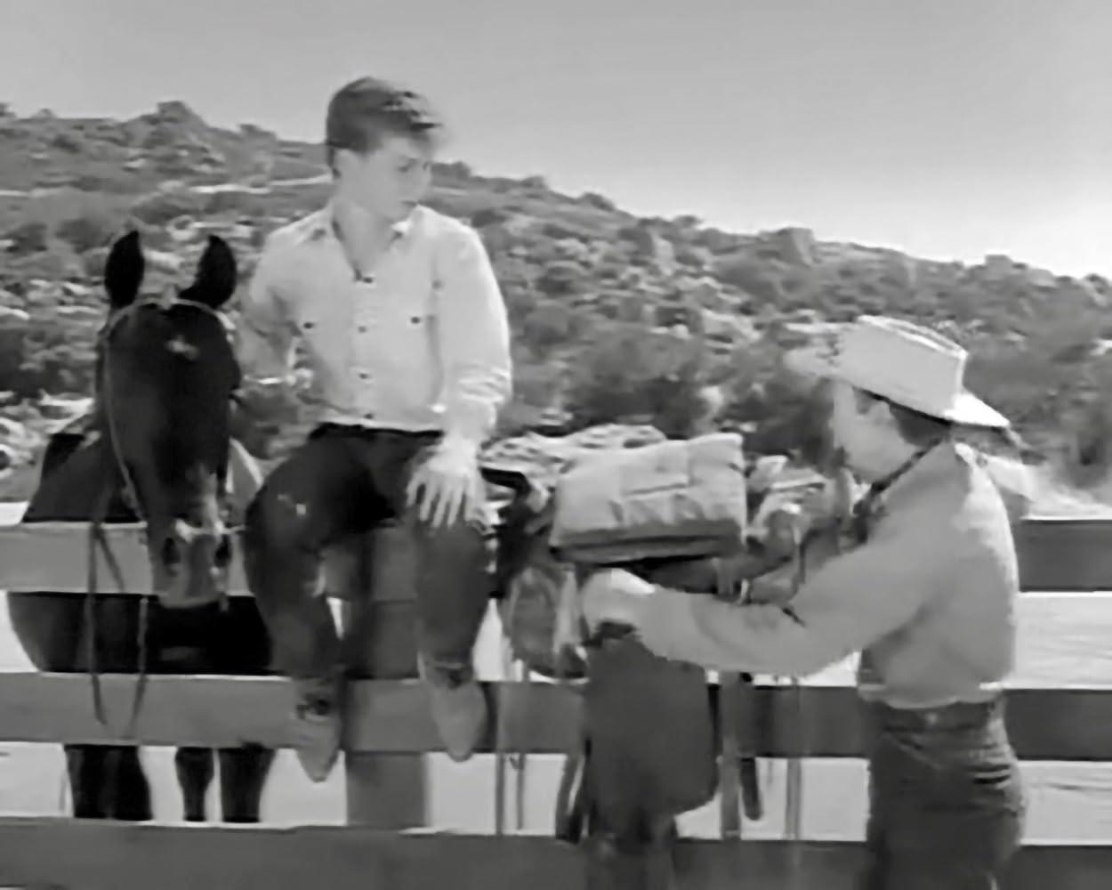 Zwei Jungen in Westernkleidung und ein Pferd an einem Zaun. Ein Junge sitzt, einer verstellt Satteltaschen.