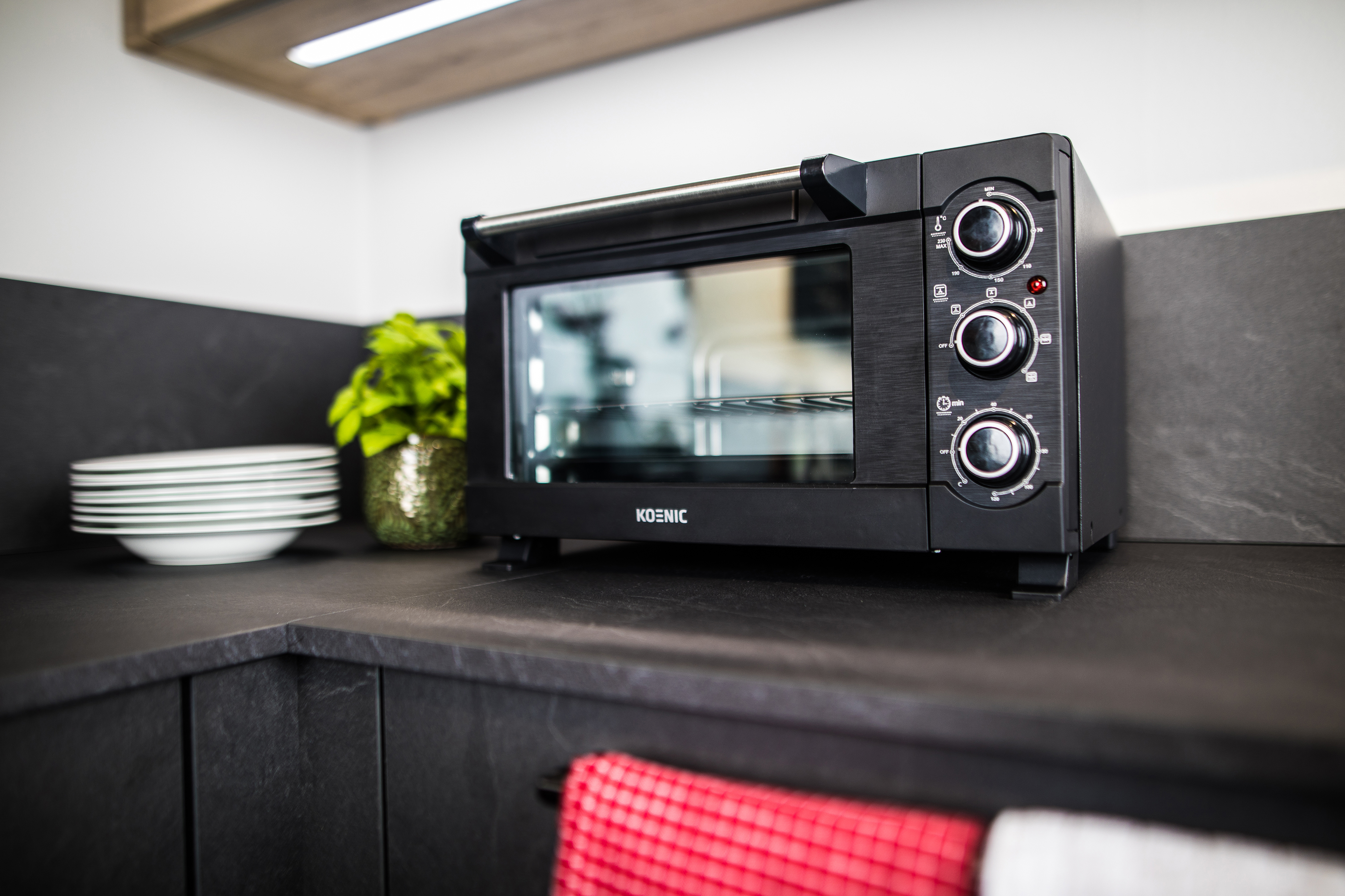 Czarny mini piekarnik Koenic na ciemnym blacie z talerzami i rośliną.
