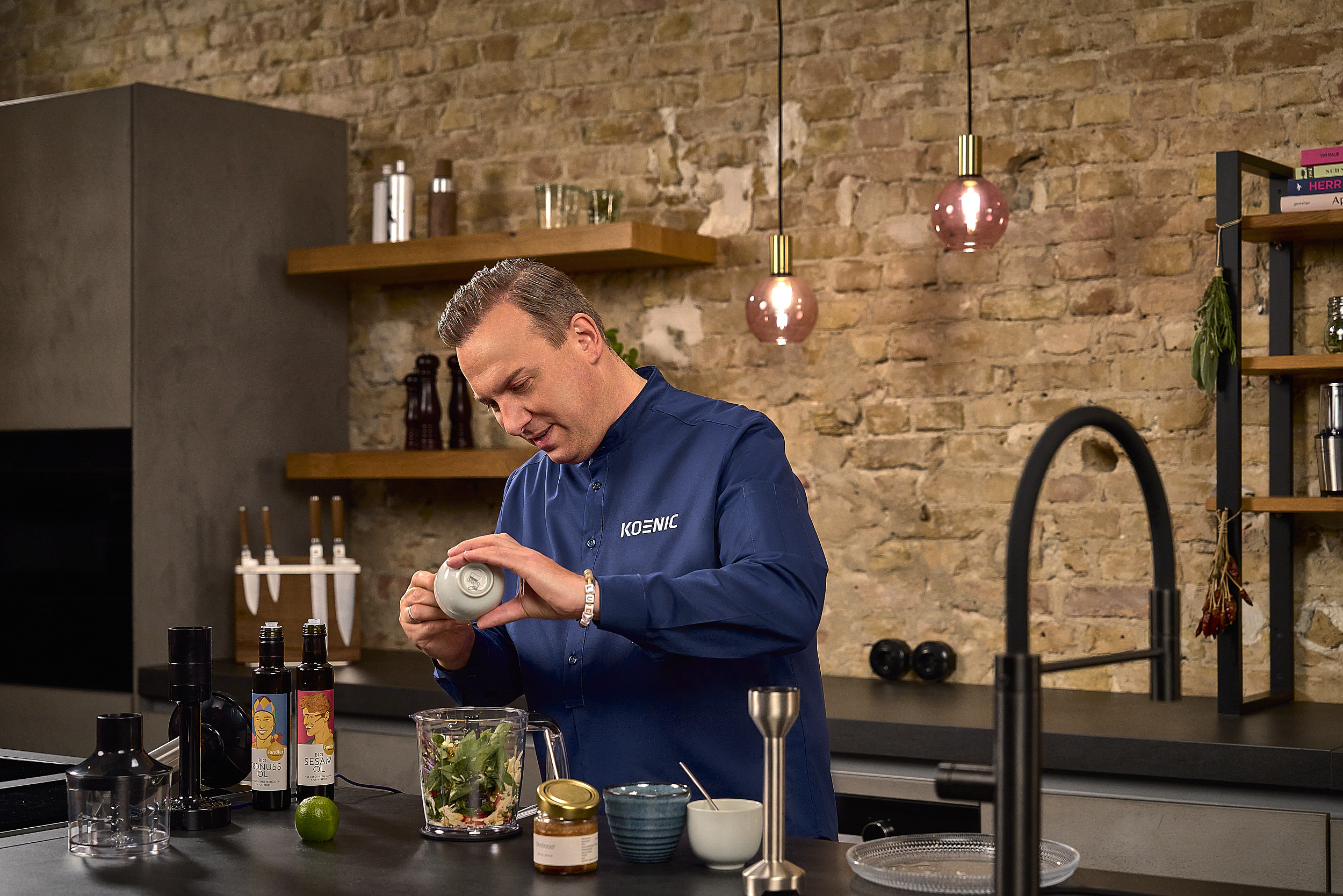 Mężczyzna w niebieskiej koszuli KOENIC dodaje składniki do blendera w kuchni.