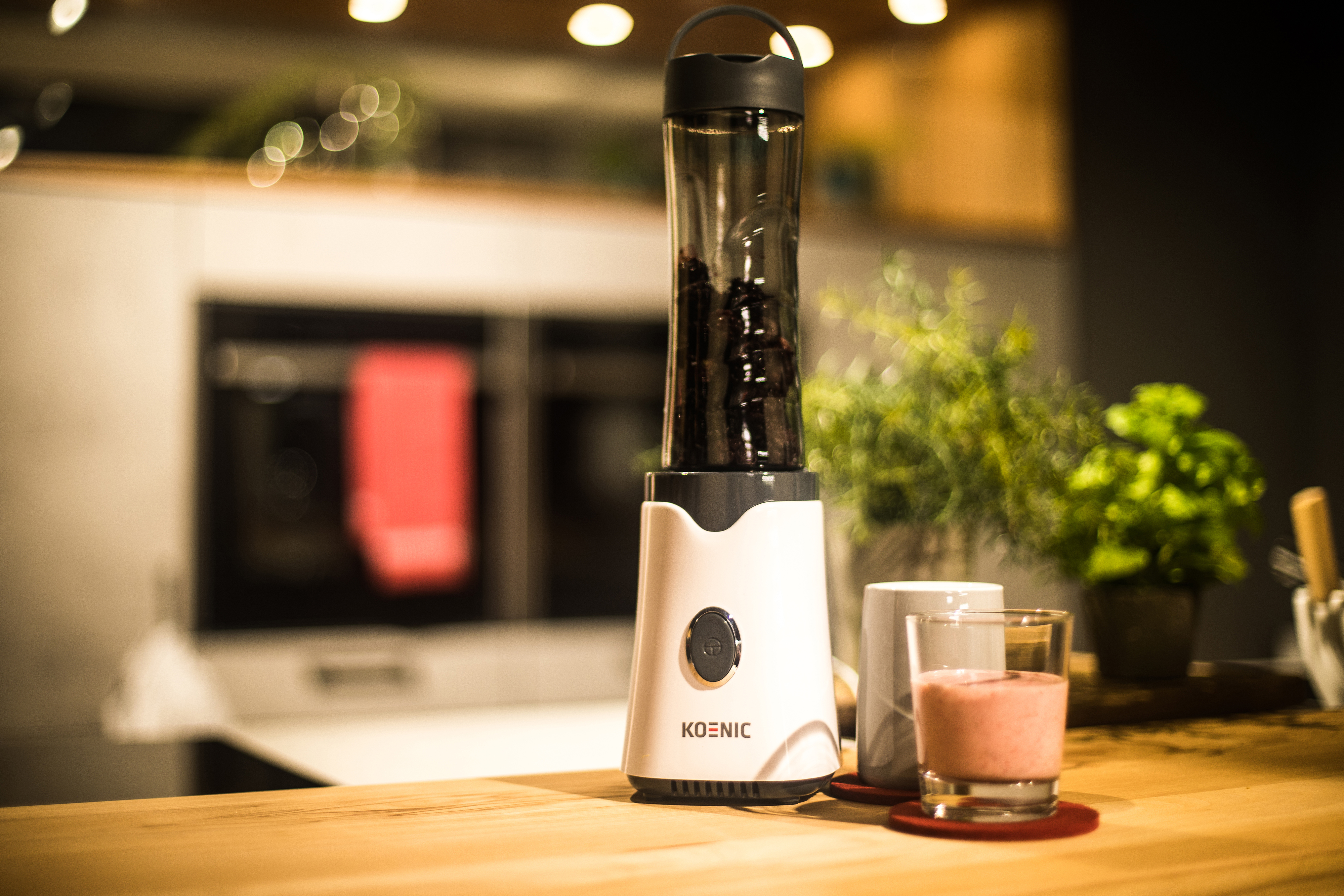 Biały blender z czarną pokrywką obok szklanki smoothie na drewnianym stole.