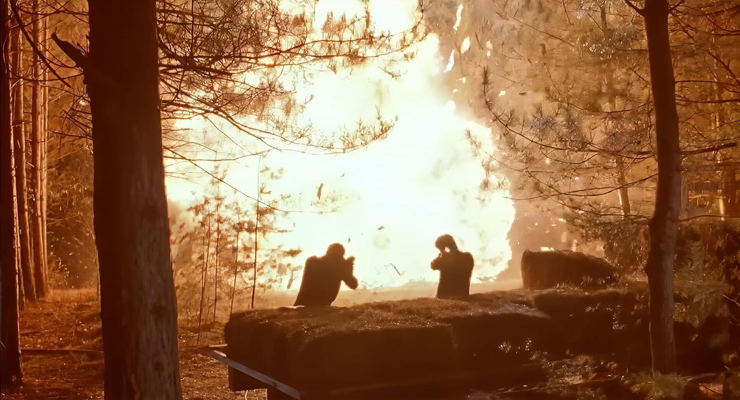 Zwei Silhouetten in einem Wald mit einer großen Explosion im Hintergrund.