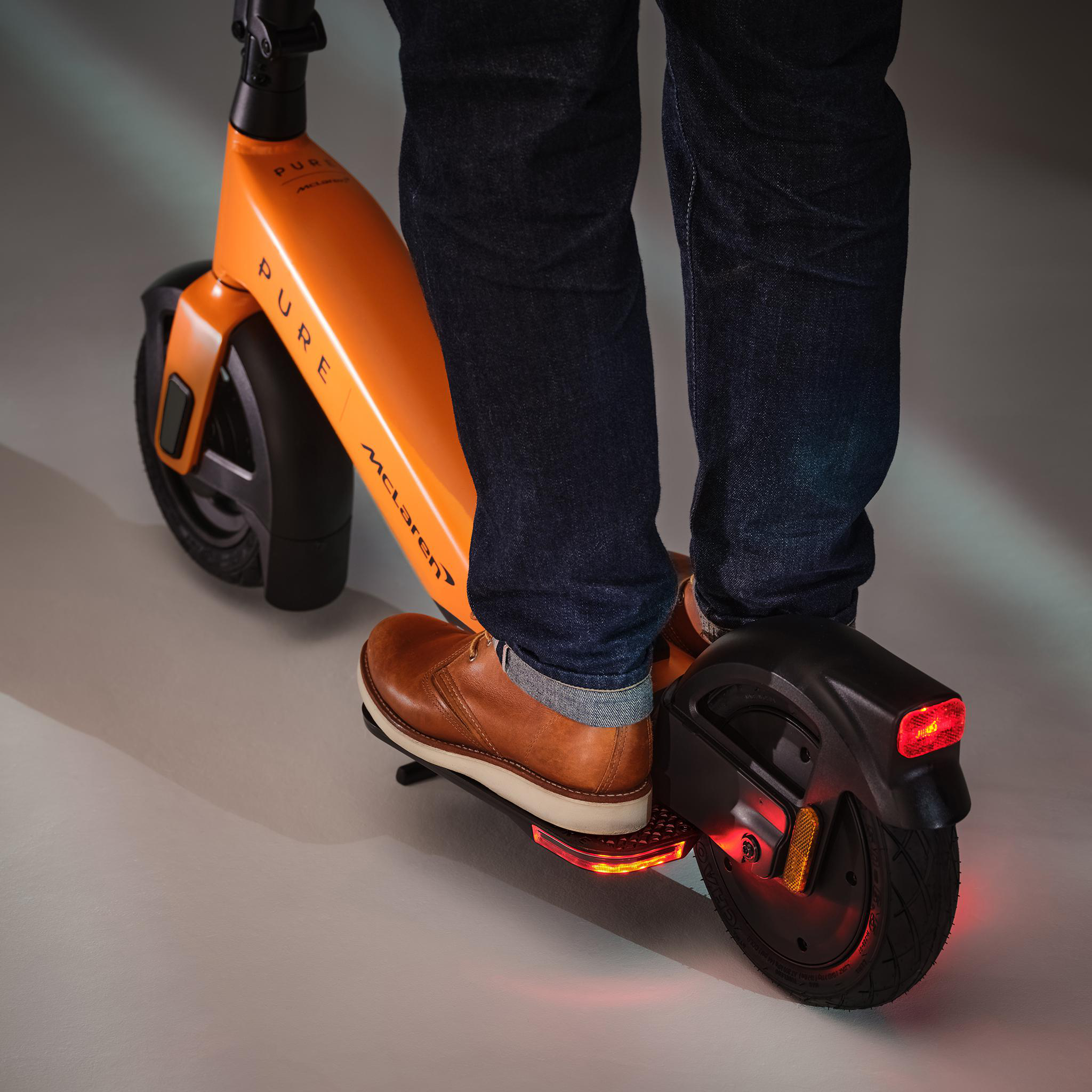 Eine Person steht auf einem orangefarbenen E-Scooter mit Jeans und braunen Schuhen.