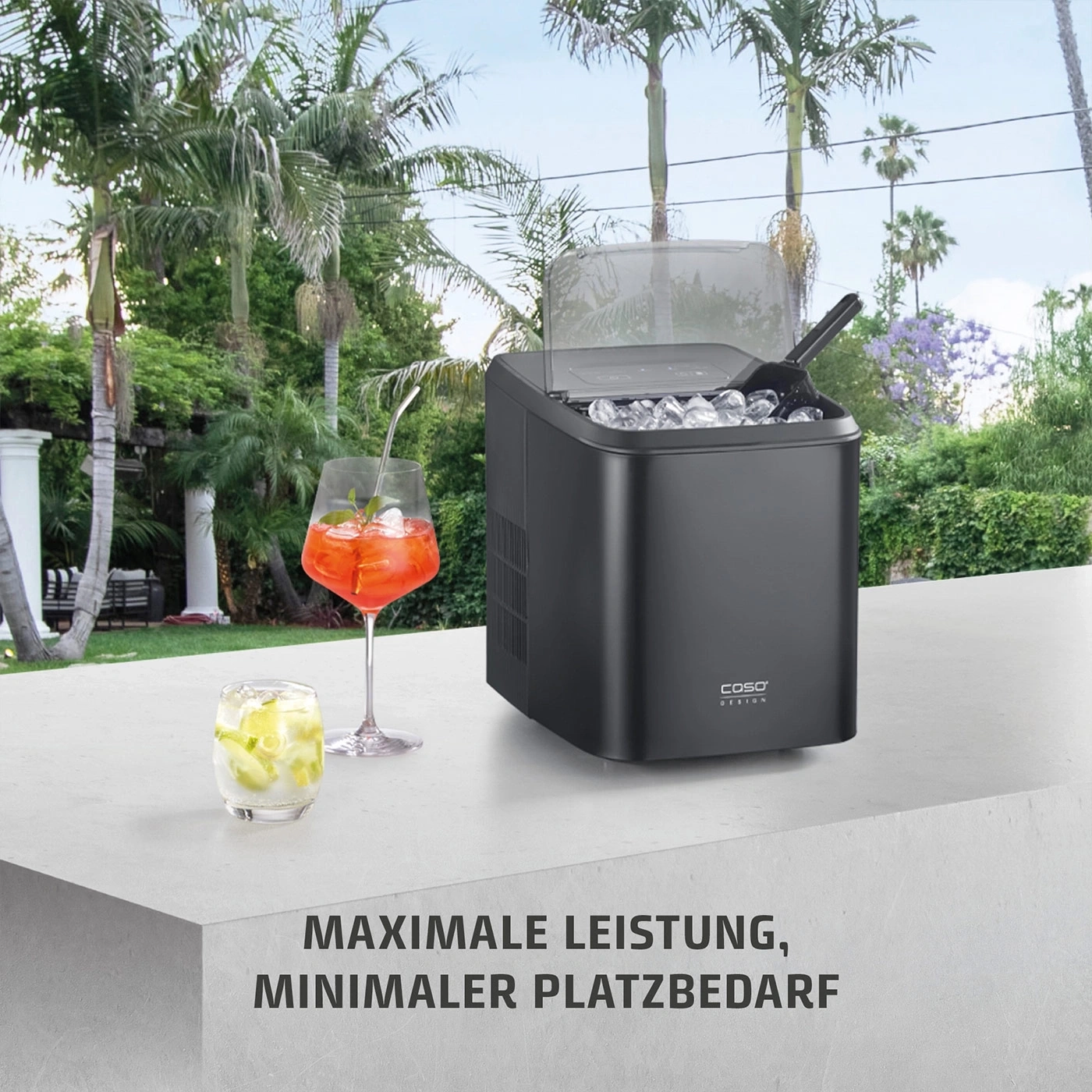 Ein schwarzer Eiswürfelbereiter mit gefülltem Behälter, daneben zwei Cocktails im Freien.