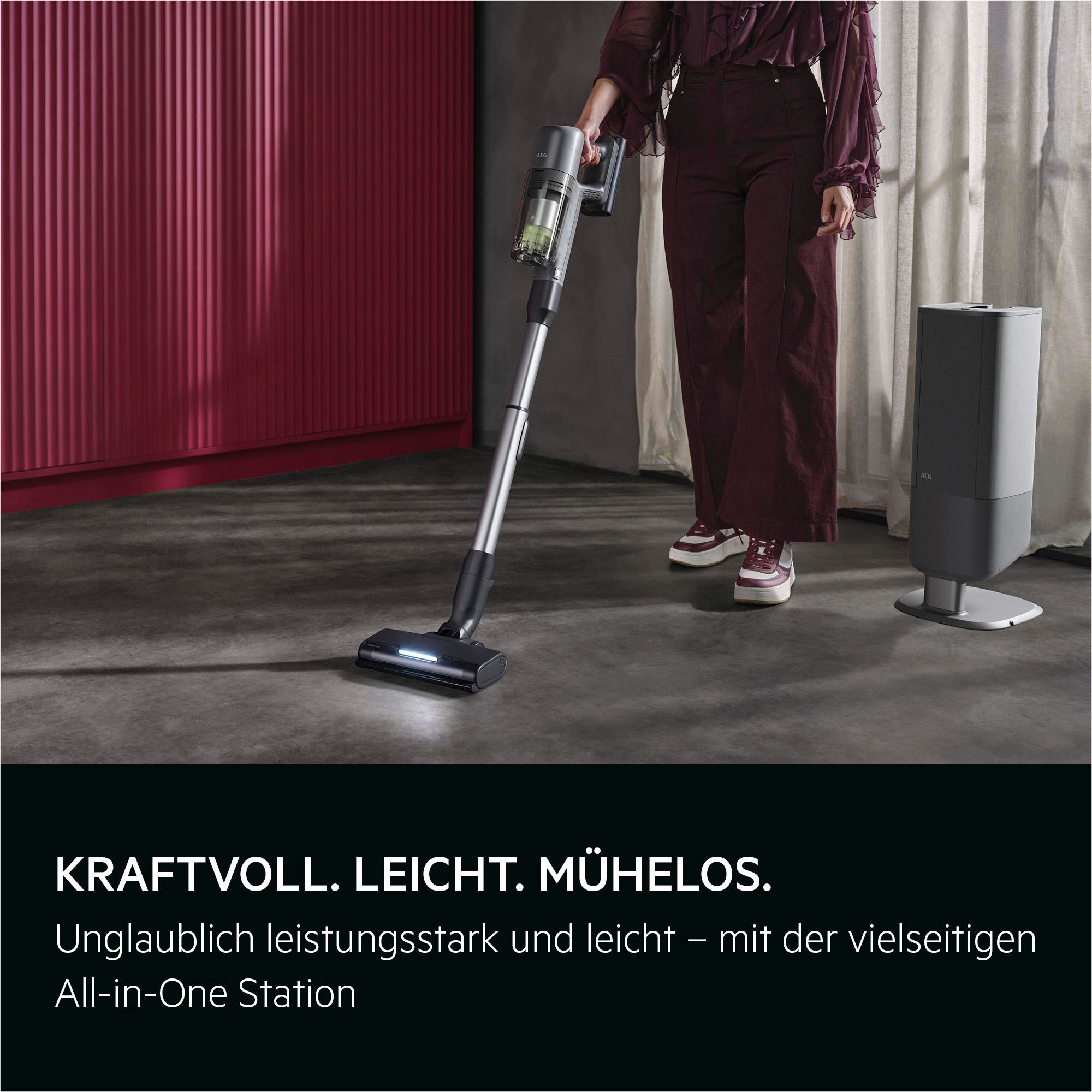 Eine Person hält einen grauen Staubsauger mit blauem Licht. Ein Lade-Stand ist rechts.