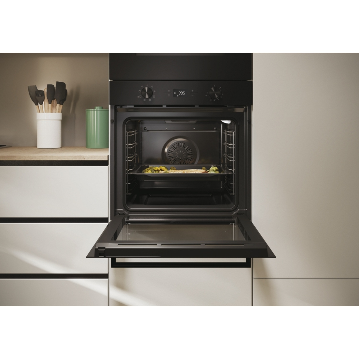 Forno nero aperto con cibo all'interno, utensili da cucina accanto.