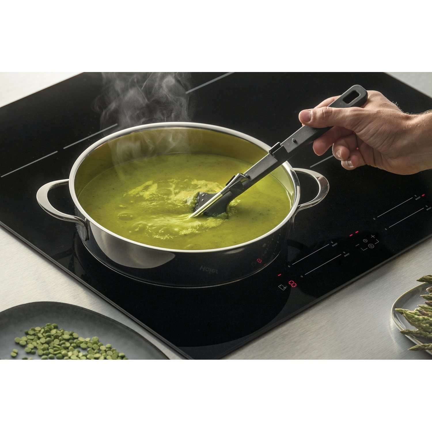 Una mano mescola zuppa verde in una pentola su un piano cottura a induzione. Piselli secchi e asparagi vicini.