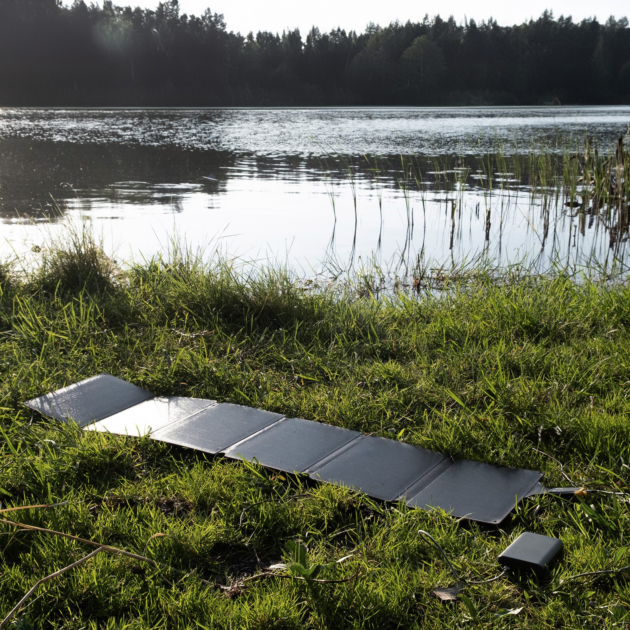 Ein Solarpanel liegt auf Gras neben einem See mit Bäumen im Hintergrund.