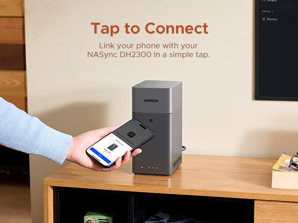 Eine Hand hält ein Smartphone neben einem grauen NAS-Gerät auf einem Holzschrank.