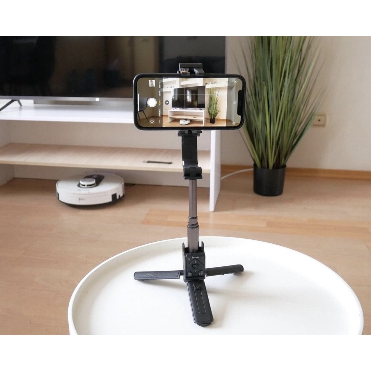 Uno smartphone su un treppiede gimbal su un tavolo, con un robot aspirapolvere sullo sfondo.