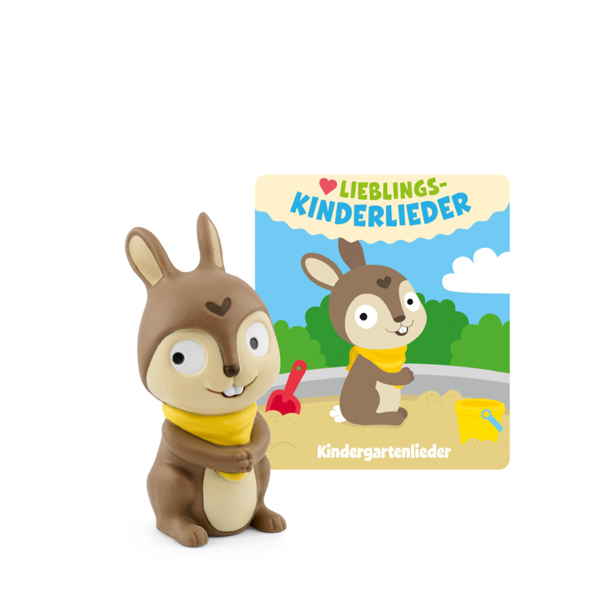 Braunes Kaninchen im Sand mit Schaufel und Eimer, im Hintergrund grüne Bäume.