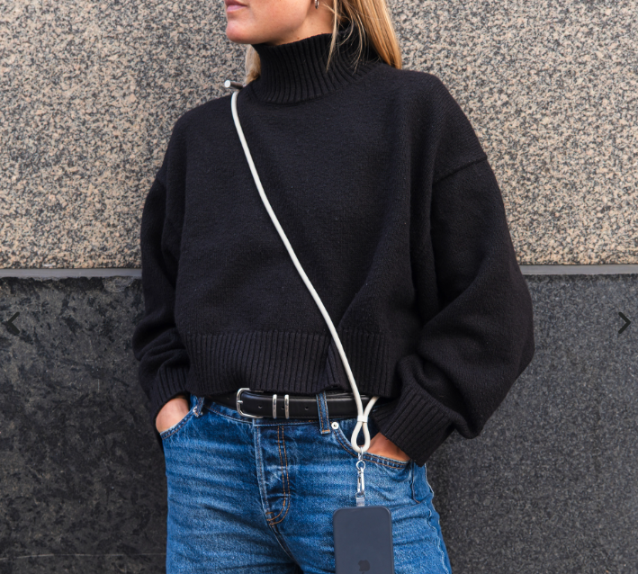 Frau in schwarzem Rollkragenpullover und blauer Jeans mit Handy-Schlinge, steht vor Steinwand.