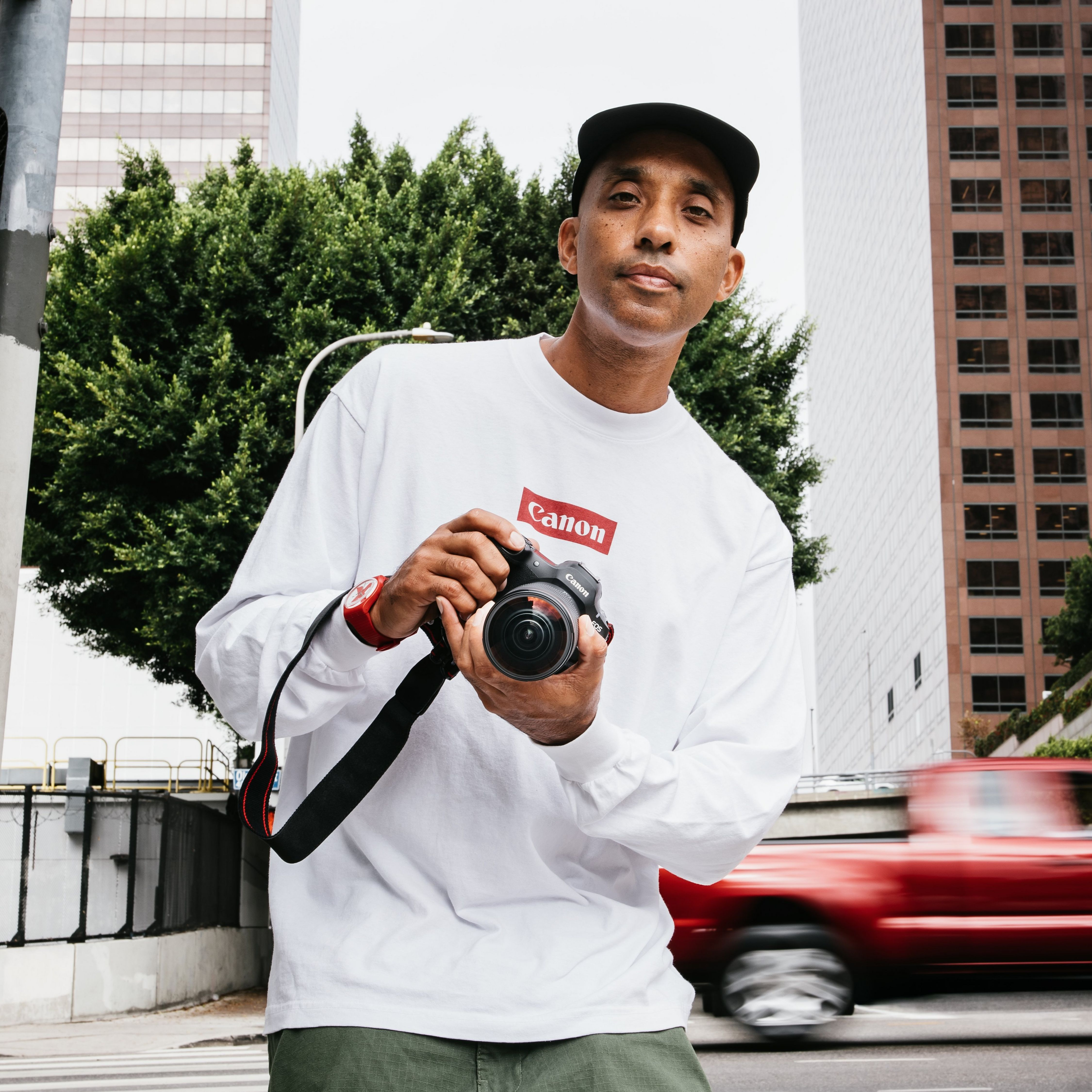 Homme avec appareil photo devant des immeubles, voiture rouge passe.