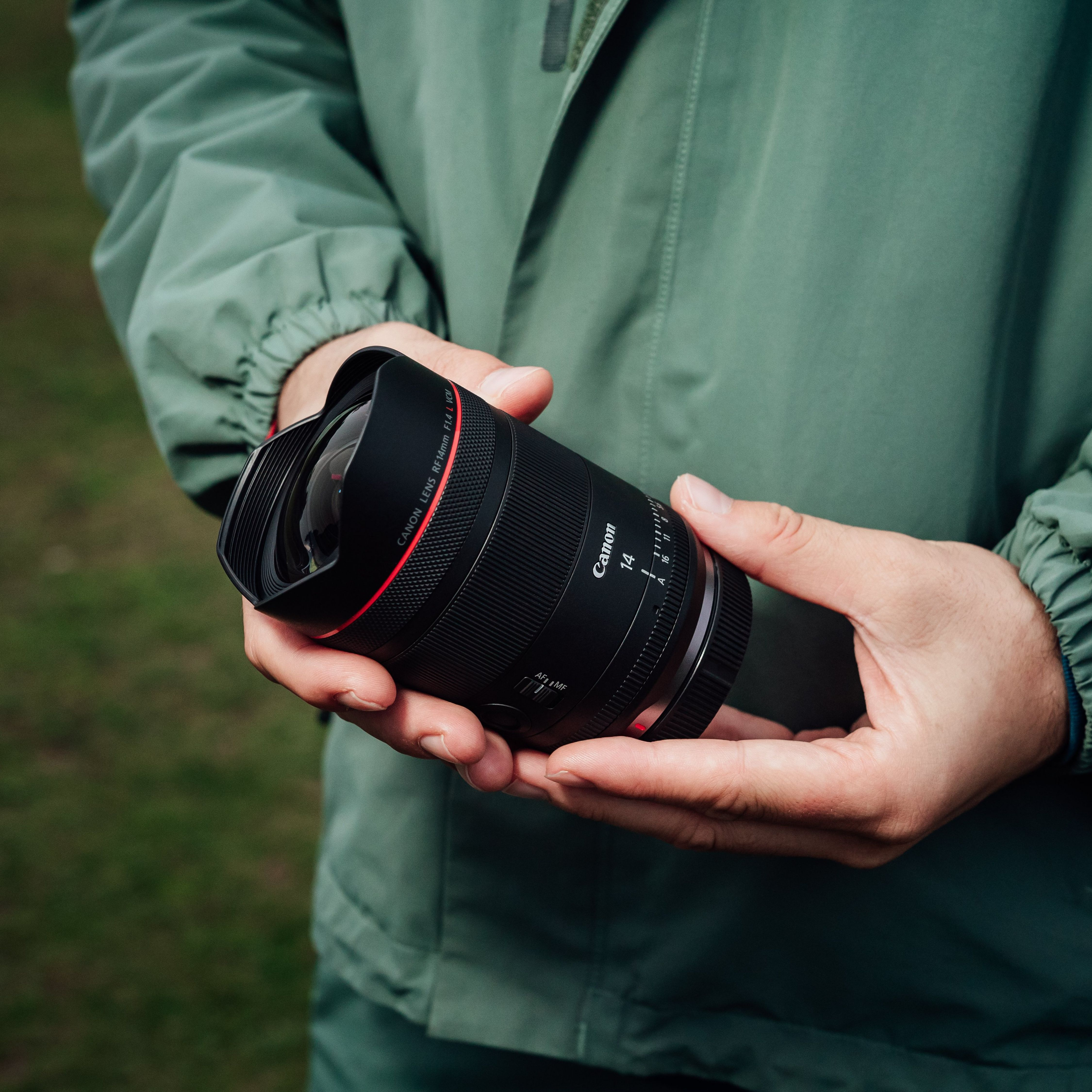 Personne tenant un objectif Canon RF 14mm F1.4 L VCM noir, fond vert.