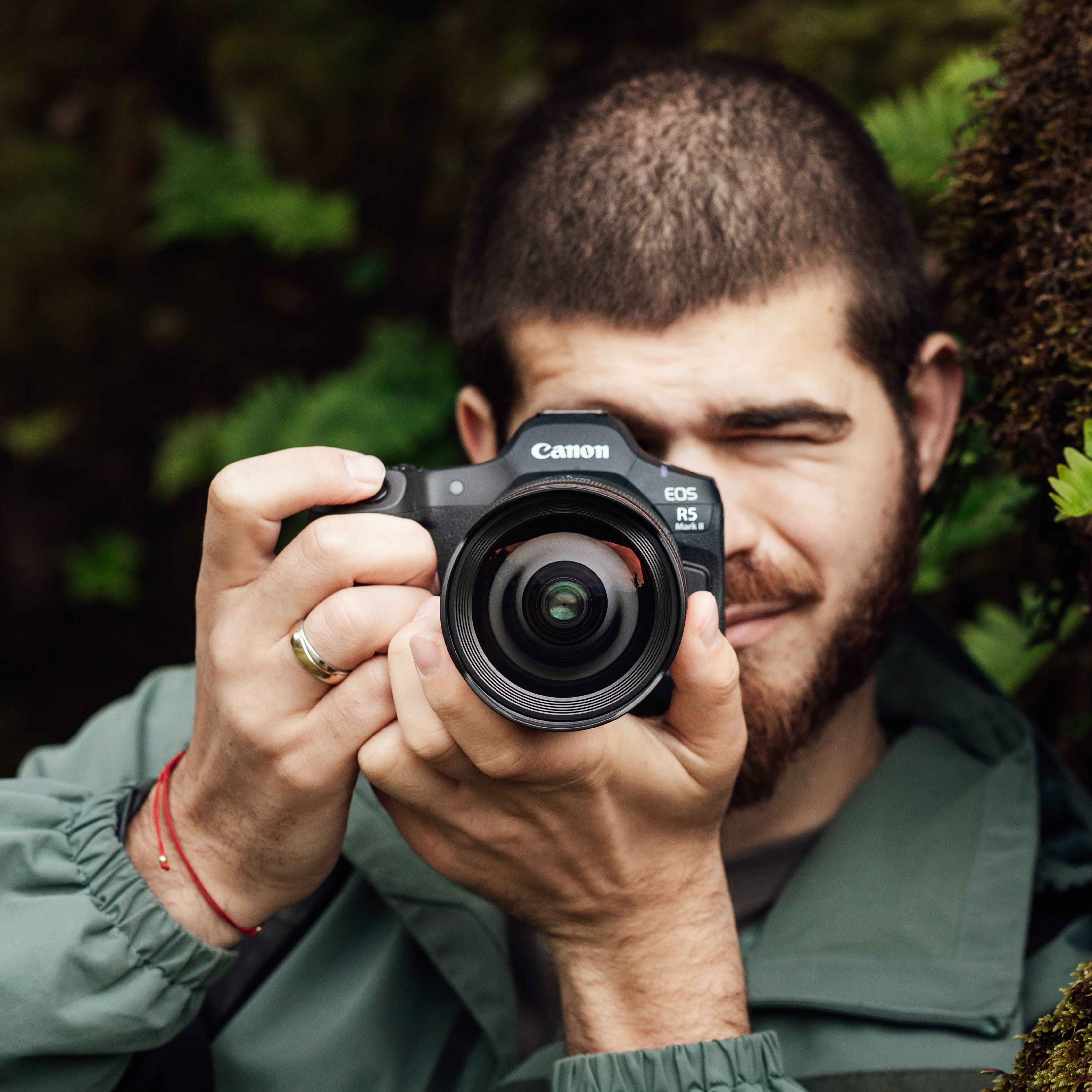 Un homme barbu tient un appareil photo Canon devant son visage, en extérieur.